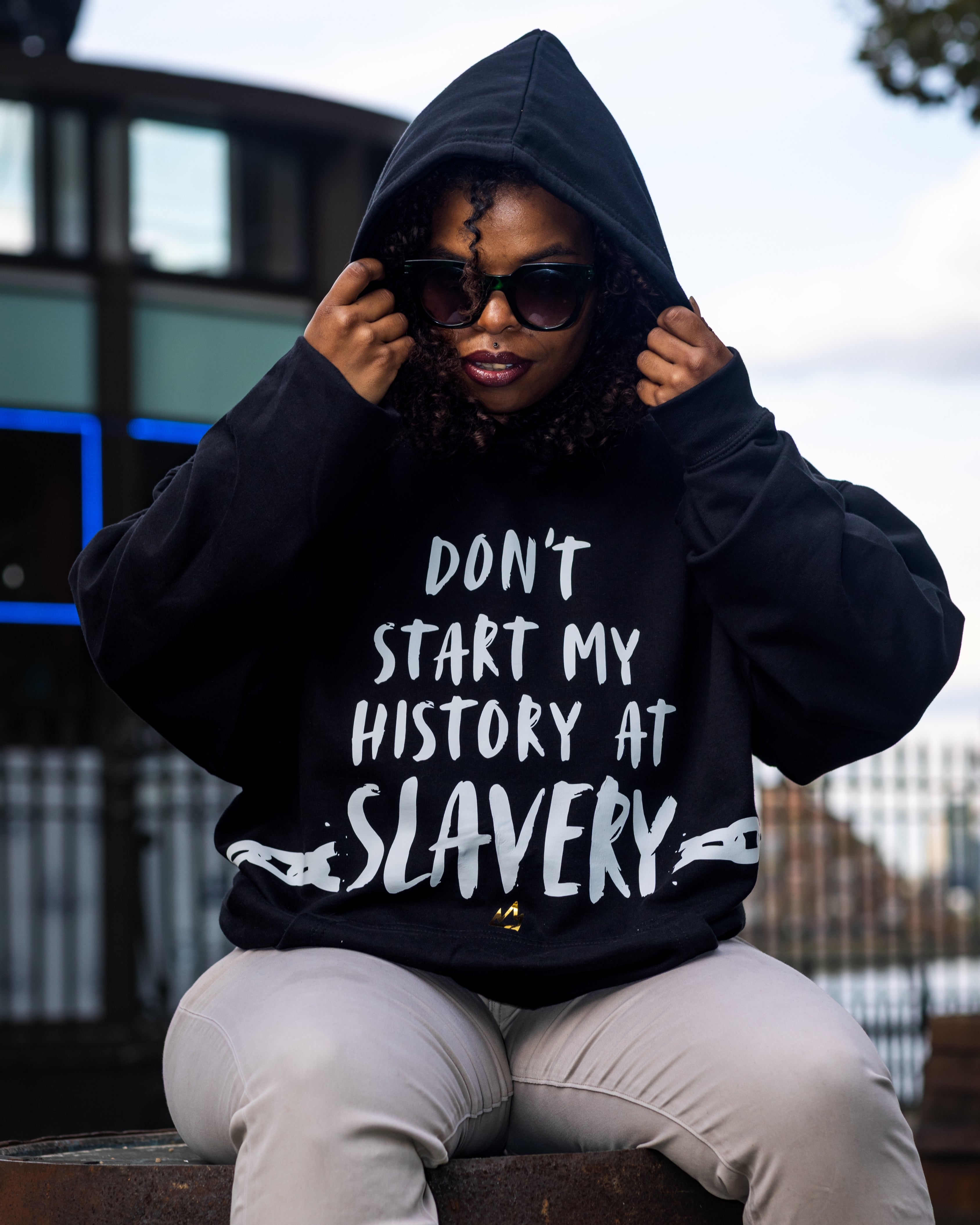 Model wearing a black hoodie with don't start my history at slavery sitting outdoors.